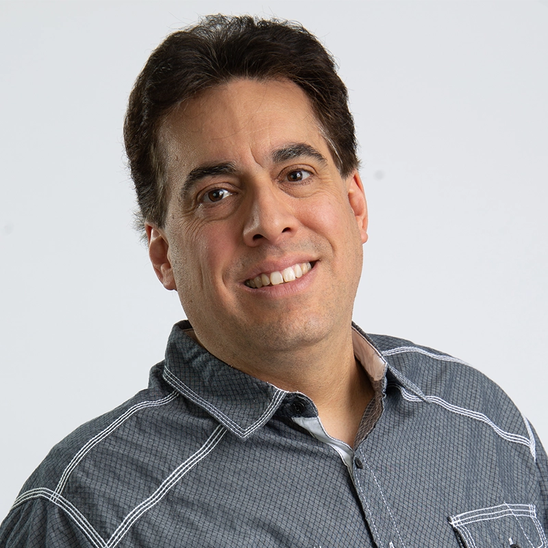Eric Scott Klein, a man with dark hair and light skin, smiles at the camera. He is wearing a gray button-up shirt with white stitching over a beige undershirt, set against a plain white background.