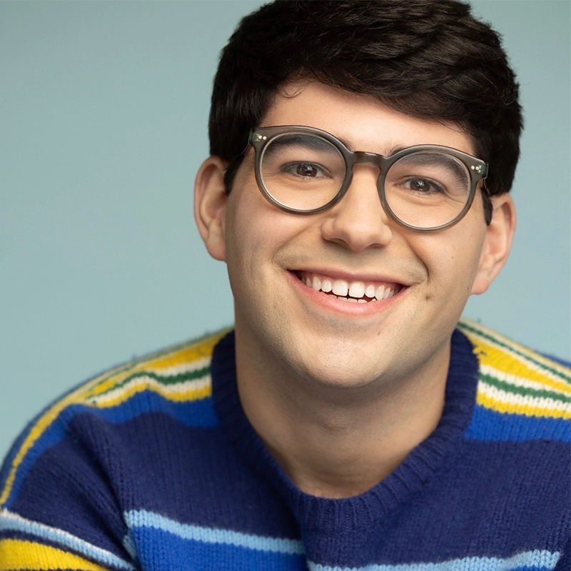 A smiling young adult with short dark hair and round glasses, wearing a blue sweater with yellow, green, and white stripes, poses in front of a plain light blue background.