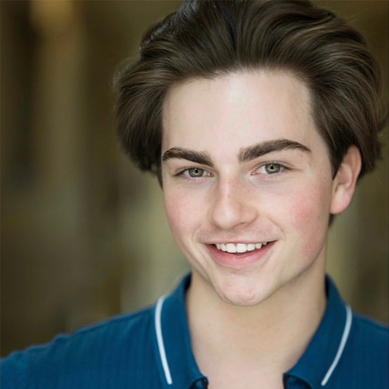A young person with fair skin and thick brown hair smiles at the camera. They wear a blue collared shirt with white trim. The background is softly blurred, focusing attention on their friendly expression and clear complexion.