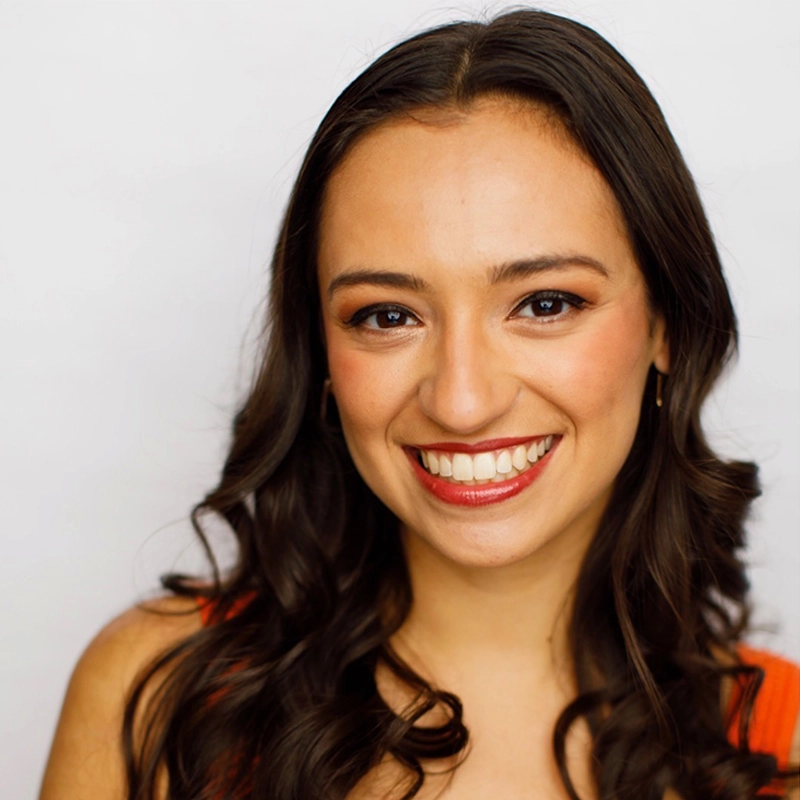 A young woman with long, wavy dark hair smiles warmly at the camera. She is wearing a sleeveless orange top and small hoop earrings. Her makeup is natural, and the background is plain white, drawing focus to her cheerful expression.
