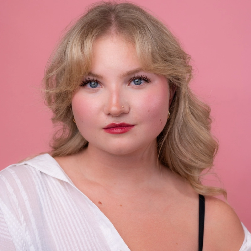 Ainsley Martell, a young woman with wavy blonde hair and blue eyes, poses against a pink background. She wears a white, slightly sheer top over a black strap and has natural makeup with rosy cheeks and red lipstick, looking confidently at the camera.