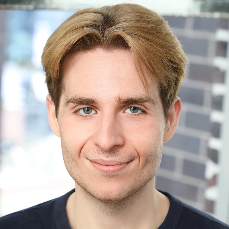 A young man with light skin and short, parted blond hair smiles slightly at the camera. He has blue eyes and stubble. He wears a dark crew-neck shirt. The background is blurred, showing brickwork and daylight through a window—an inviting portrait of Matt Tierney in a candid moment.