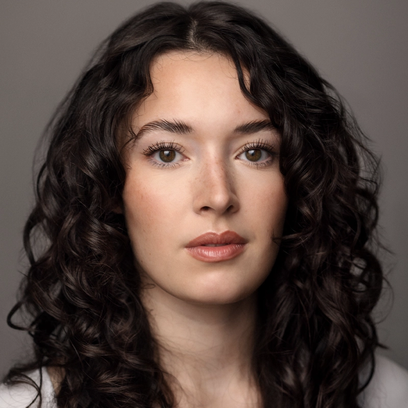 A young woman with long, dark, curly hair, clear skin, and brown eyes looks directly at the camera. She has natural makeup and neutral lips, and she is set against a plain, soft gray background.