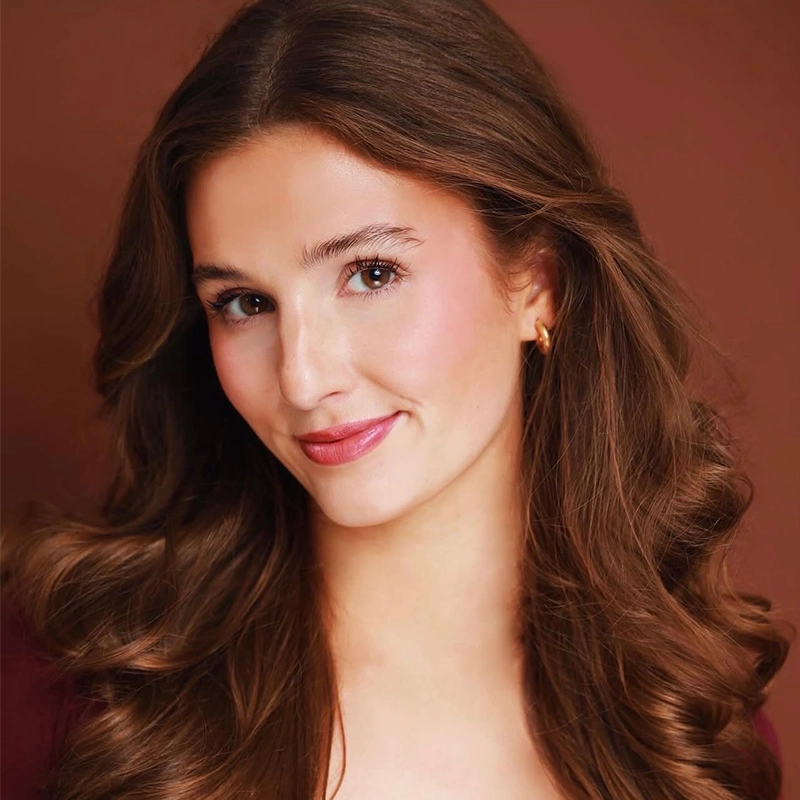 A young woman with long, wavy brown hair poses against a warm brown background. She is smiling softly, has light skin, and wears subtle makeup. She is dressed in a burgundy top and small gold hoop earrings, giving a natural and elegant appearance.