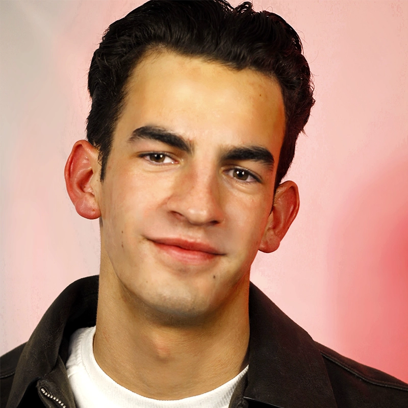 A young man with short dark hair, thick eyebrows, and light skin smiles softly. He wears a dark jacket over a white shirt. The blurred background has pink and beige tones, creating a warm, gentle atmosphere.