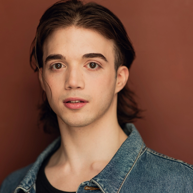 Maxwell Parmelee, a young person with fair skin, medium-length brown hair, and brown eyes, wears a blue denim jacket over a black shirt. They look directly at the camera with a neutral expression against a solid, muted brown background.