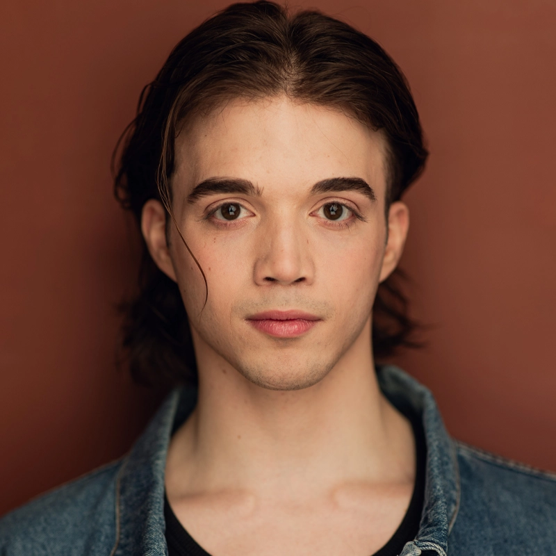 A young person with fair skin, straight brown hair, and brown eyes gazes directly at the camera. Maxwell Parmelee wears a denim jacket over a black shirt. The background is a solid, warm reddish-brown color, and the lighting is soft, emphasizing their facial features.