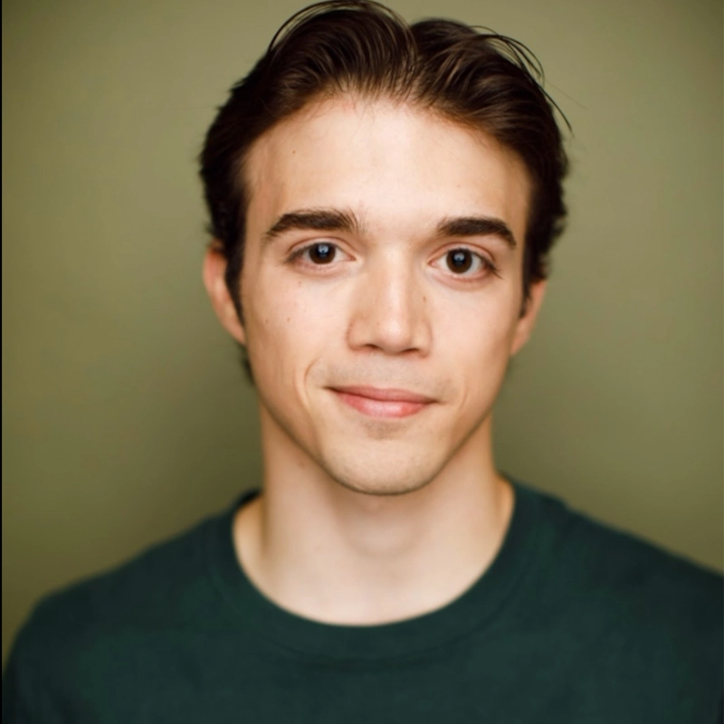 Maxwell Parmelee, a young man with fair skin, dark brown hair parted back, and brown eyes, looks at the camera with a gentle smile. He wears a dark green crew-neck shirt against a plain, muted green background.