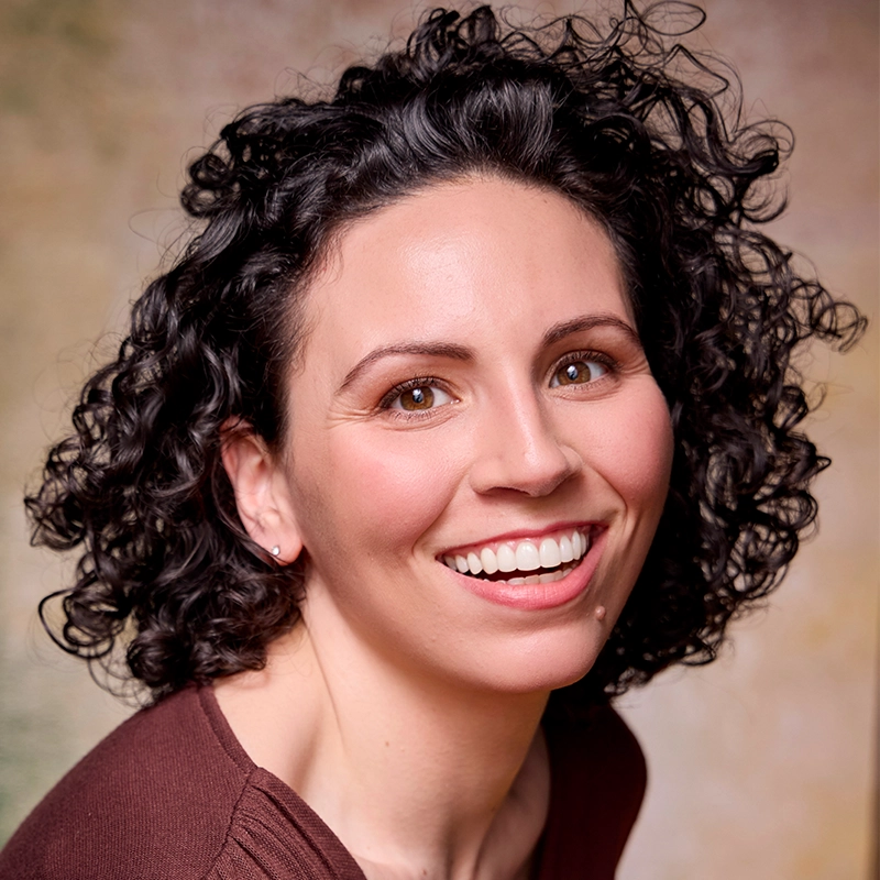 Shamiea Thompson, a woman with medium-toned skin and curly dark hair, smiles brightly at the camera. She wears a maroon top and natural makeup. The softly blurred earth-toned background highlights her cheerful expression.