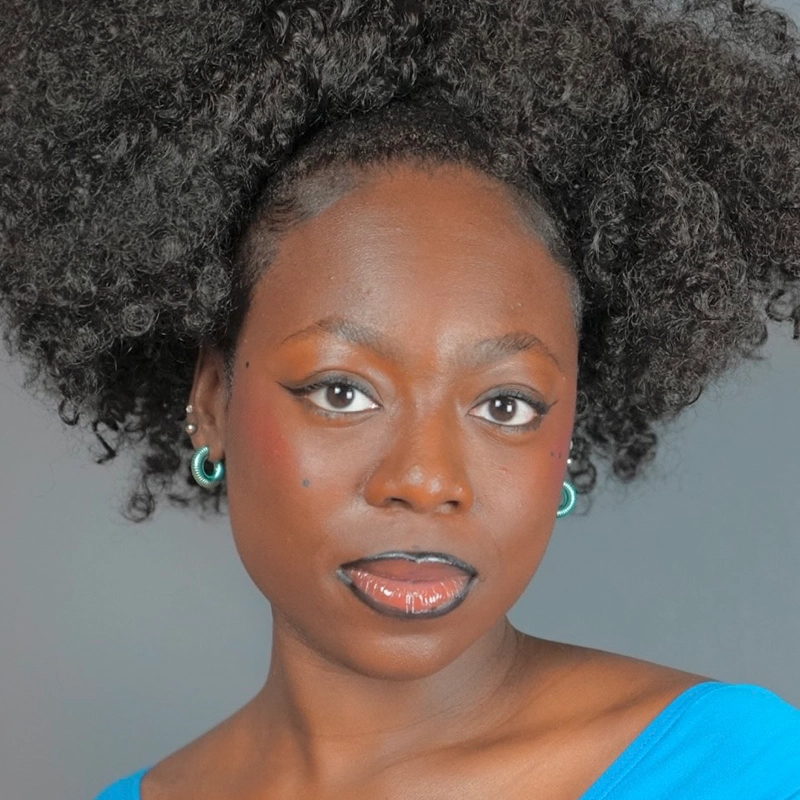 Shamiea Thompson, a woman with dark skin and voluminous, natural curly hair, looks confidently at the camera. She wears turquoise hoop earrings, has winged eyeliner, glossy lips, and a blue top. A plain background highlights her expressive face.