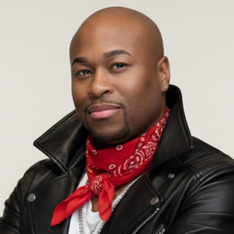 A confident man with a shaved head, trimmed beard, and stud earrings—Eric Scott Klein—wears a black leather jacket, white shirt, and red bandana around his neck. He faces forward with a slight smile against a plain, light-colored background.