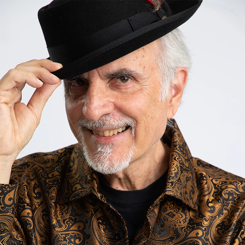 Eric Scott Klein, an older man with a goatee and mustache, smiles as he tilts a black fedora with one hand. He wears a brown paisley-patterned shirt over a black shirt, set against a plain white background.