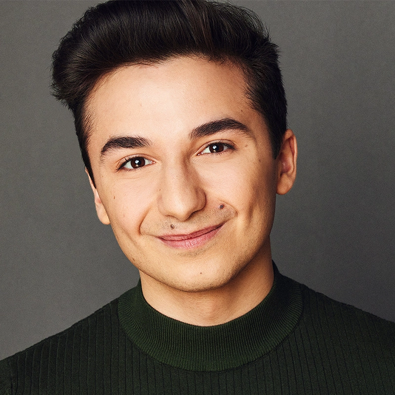 A young person with short dark hair, clear skin, and a faint mustache smiles gently at the camera. Wes D'Alelio wears a dark green, ribbed, high-neck sweater and is posed in front of a plain gray background.