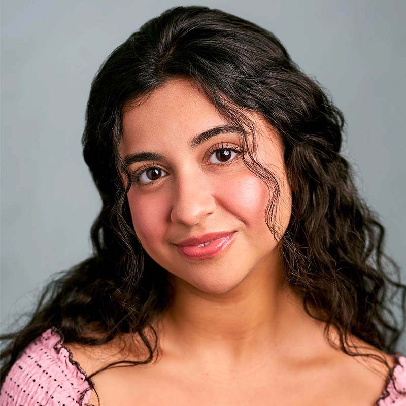A young woman with long, wavy dark hair and warm-toned skin smiles softly. Camila Serrano wears a light pink top and natural makeup, with loose curls framing her face. The background is plain and softly lit in light gray.