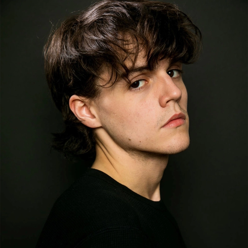 A young person with wavy brown hair and fair skin looks seriously at the camera over their shoulder. They wear a black top and stand against a dark background, with soft lighting highlighting their facial features—reminiscent of Anthony Chavers.