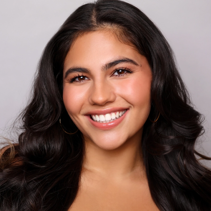 A close-up portrait of a smiling young woman with long, wavy dark hair and brown eyes. She wears simple gold hoop earrings against a plain, light grey background.