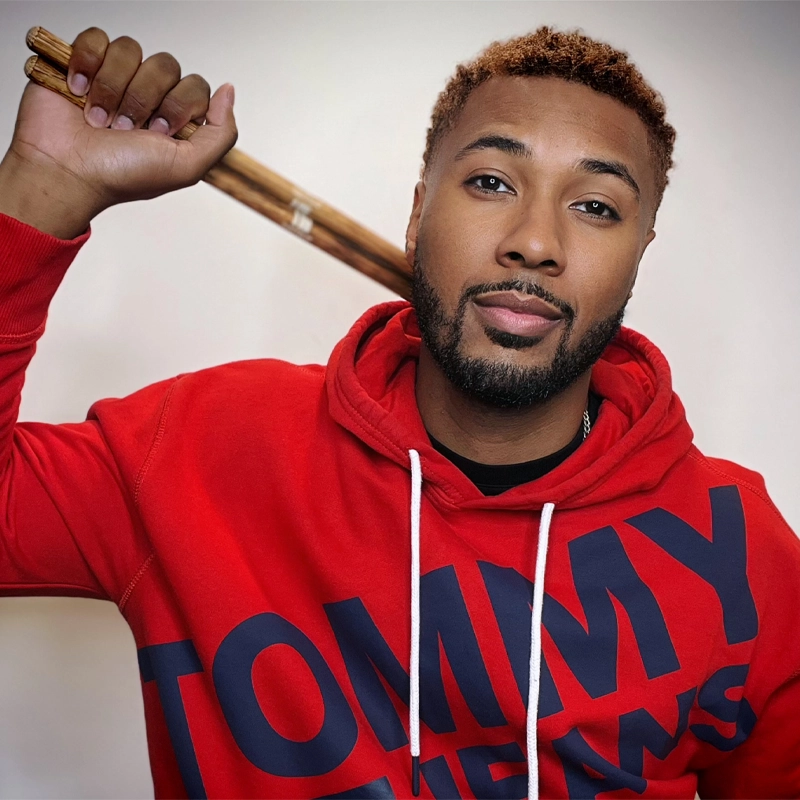 Shawn Dustin, with short curly hair and a trimmed beard, wears a bright red Tommy Jeans hoodie. Holding a wooden baseball bat on his shoulder, he looks confidently at the camera against a plain white background.