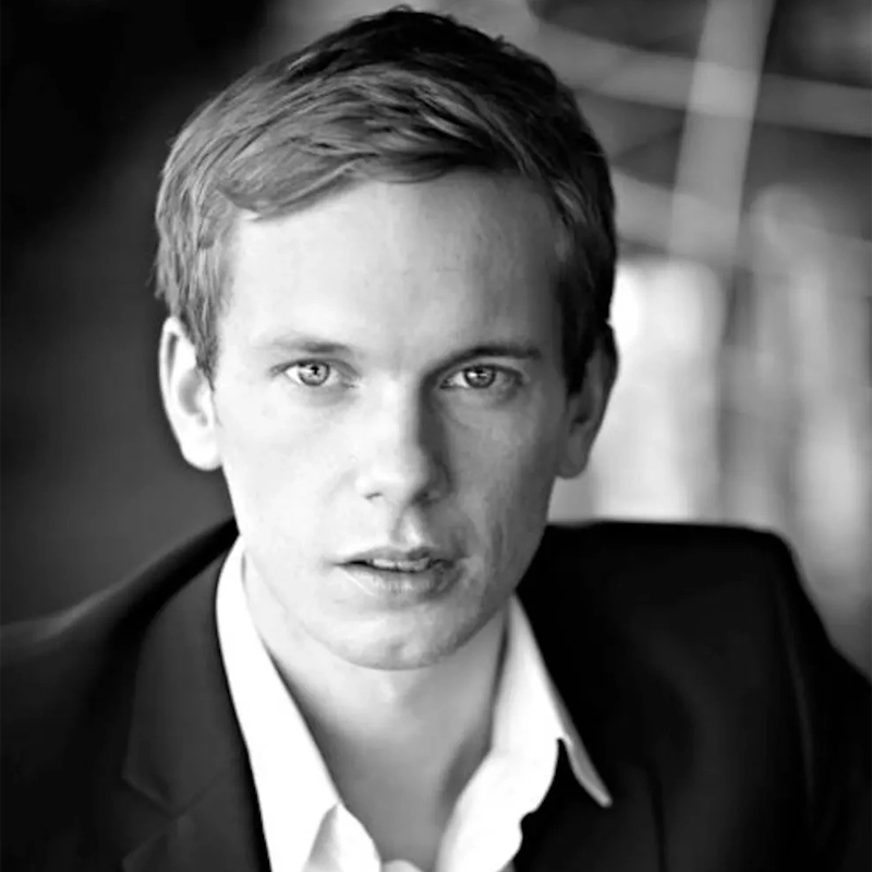 A young man with light skin, short light brown hair, and light-colored eyes looks directly at the camera. He wears a dark suit jacket over a white dress shirt. The photo is in black and white, with a softly blurred background.