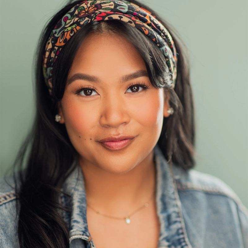 A woman with long dark hair wears a colorful patterned headband, denim jacket, and delicate necklace. She has a subtle smile, natural makeup, and small stud earrings. The background is a muted, plain color, highlighting her face and giving the portrait of Sierra Fermin a timeless elegance.