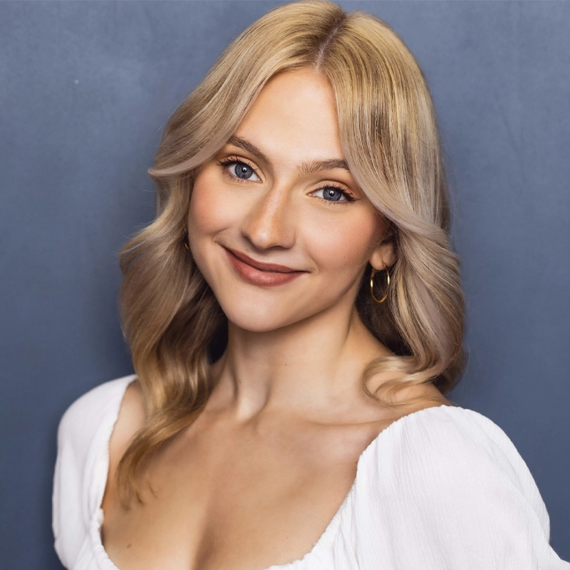 Tori Heinlein, a young woman with long, wavy blonde hair and fair skin, smiles gently. She is wearing a white, square-neck blouse and gold hoop earrings. The solid slate blue background and soft lighting highlight her features.