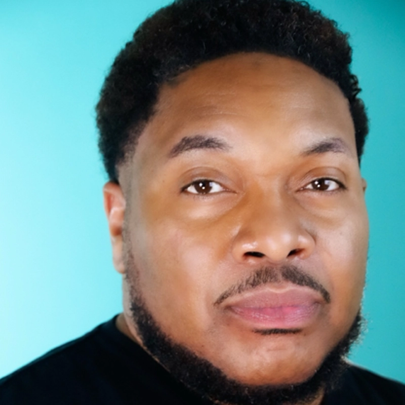 Darren Lorenzo, a man with short curly hair and a trimmed beard, looks at the camera with a neutral expression. He is wearing a black shirt against a solid light turquoise background, illuminated by even and soft lighting.