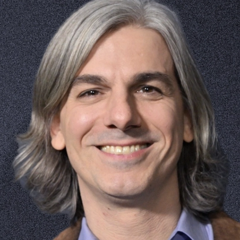 Lucas Papaelias, a smiling adult with shoulder-length, straight gray hair, wears a blue collared shirt and brown jacket, posed against a textured dark background.