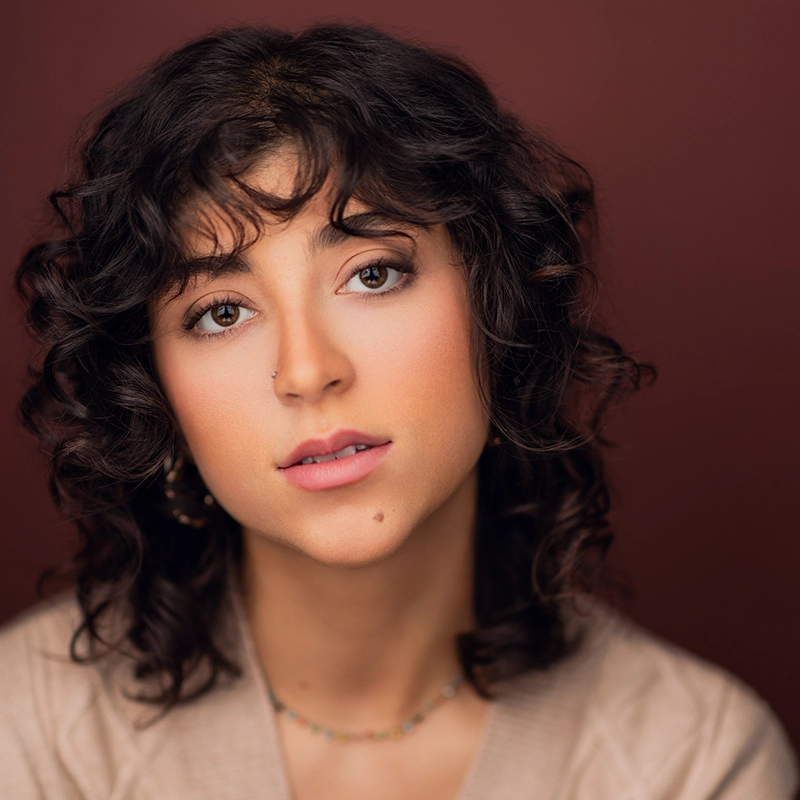 A young woman with curly dark hair and soft makeup gazes gently at the camera. She has a nose piercing, a small mole above her lip, and wears a beige top with a delicate beaded necklace. The portrait is inspired by Christian Clausnitzer’s style.