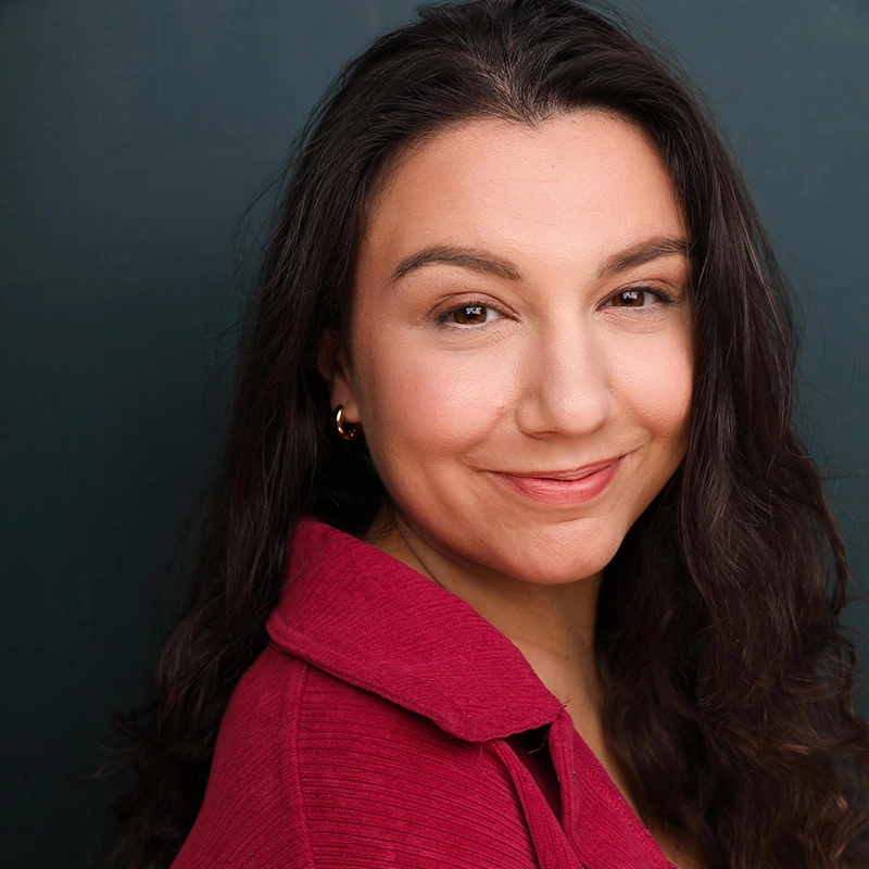 A woman with long, wavy dark hair smiles softly at the camera. Eve Tjoumakaris wears a textured, deep pink collared top and small gold hoop earrings, standing against a dark teal background, her warm expression radiating quiet confidence.
