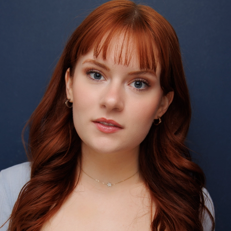 A young woman with long, wavy red hair and bangs poses in front of a dark blue background. She wears a light-colored top, small hoop earrings, and a delicate necklace with a star-shaped pendant. Her expression is neutral and her makeup is natural.