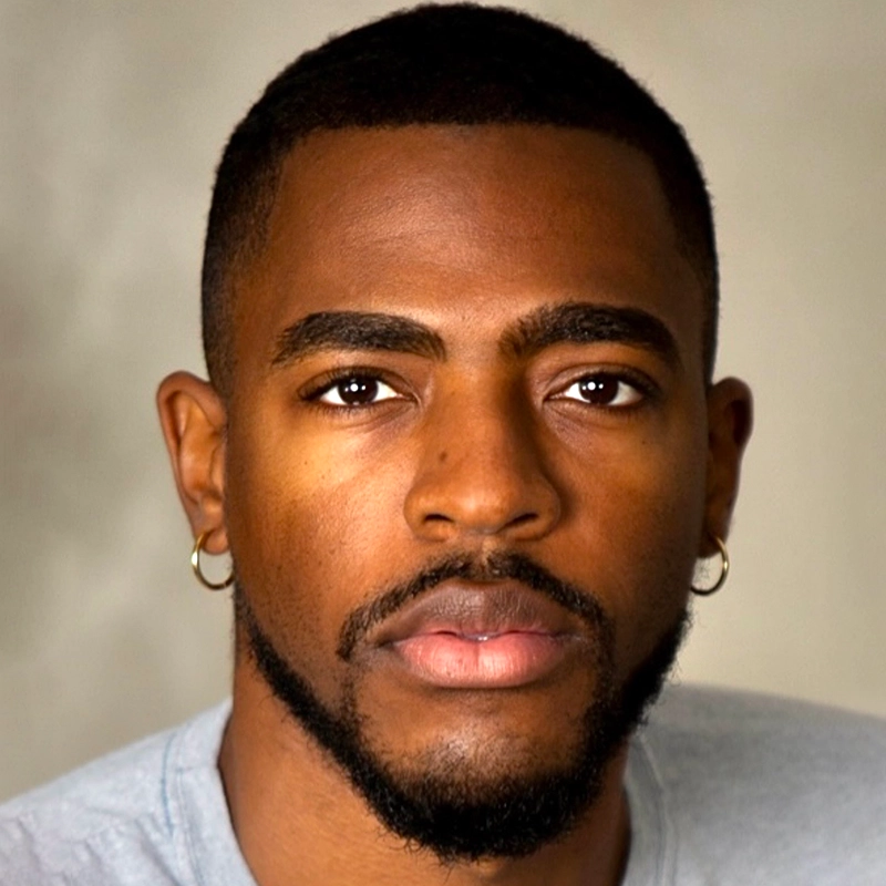 A young man with short black hair, well-groomed beard, and mustache looks directly at the camera. Christian Clausnitzer has medium brown skin, two small hoop earrings, a light gray shirt, and stands before a neutral, softly blurred background.