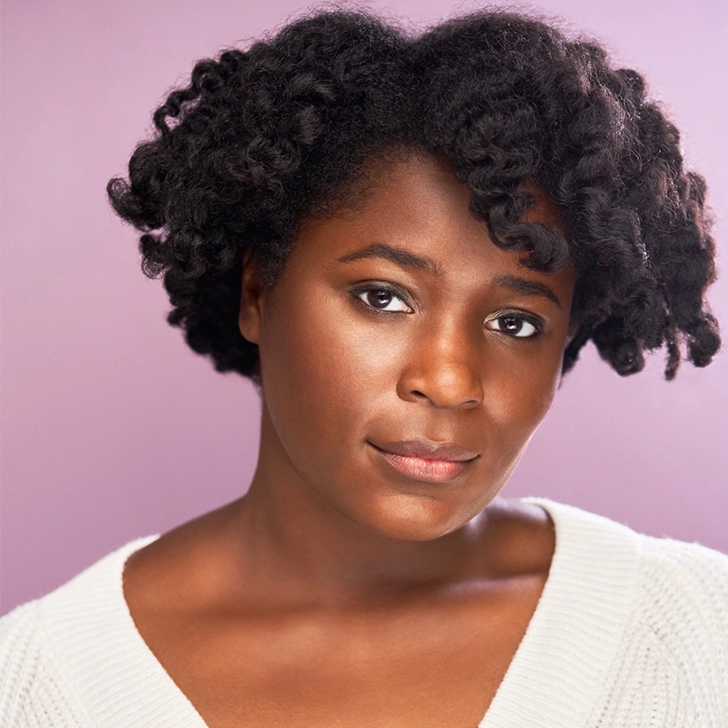 A woman with medium-dark skin and natural, textured black hair styled in loose twists looks calmly at the camera. She wears a white sweater and natural makeup, standing against a soft lavender background—an image reminiscent of Eve Tjoumakaris.