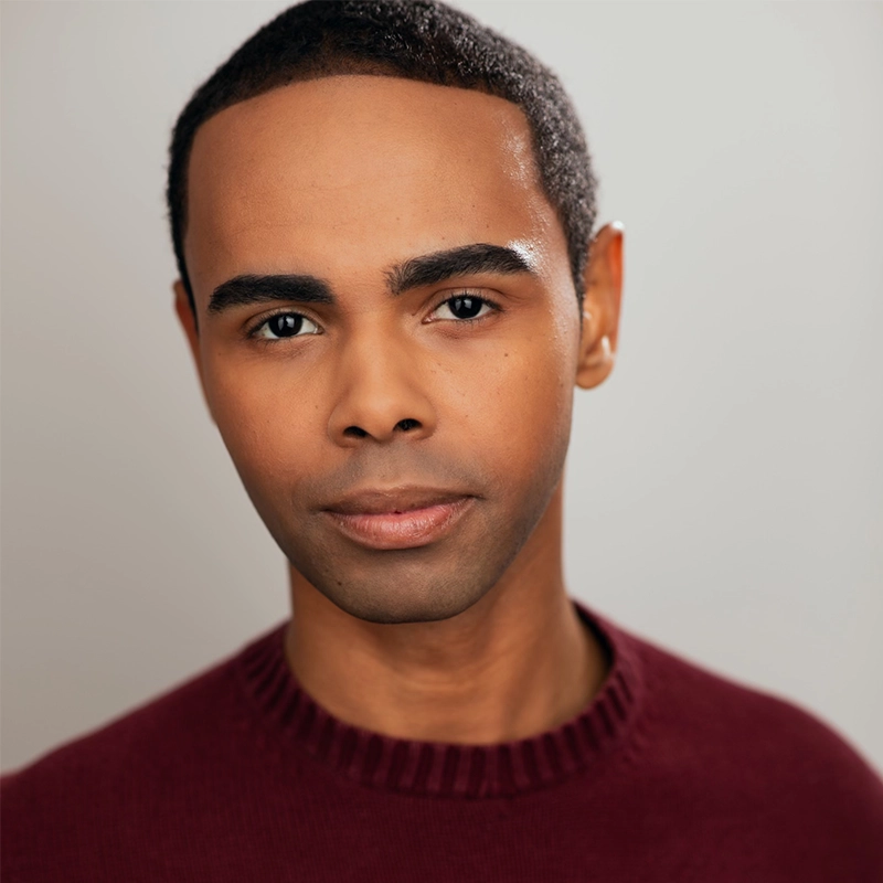 A young man with short black hair and smooth skin looks directly at the camera. Christian Clausnitzer wears a burgundy crewneck sweater and has a calm, neutral expression. The background is plain light gray, softly lit for a professional portrait effect.