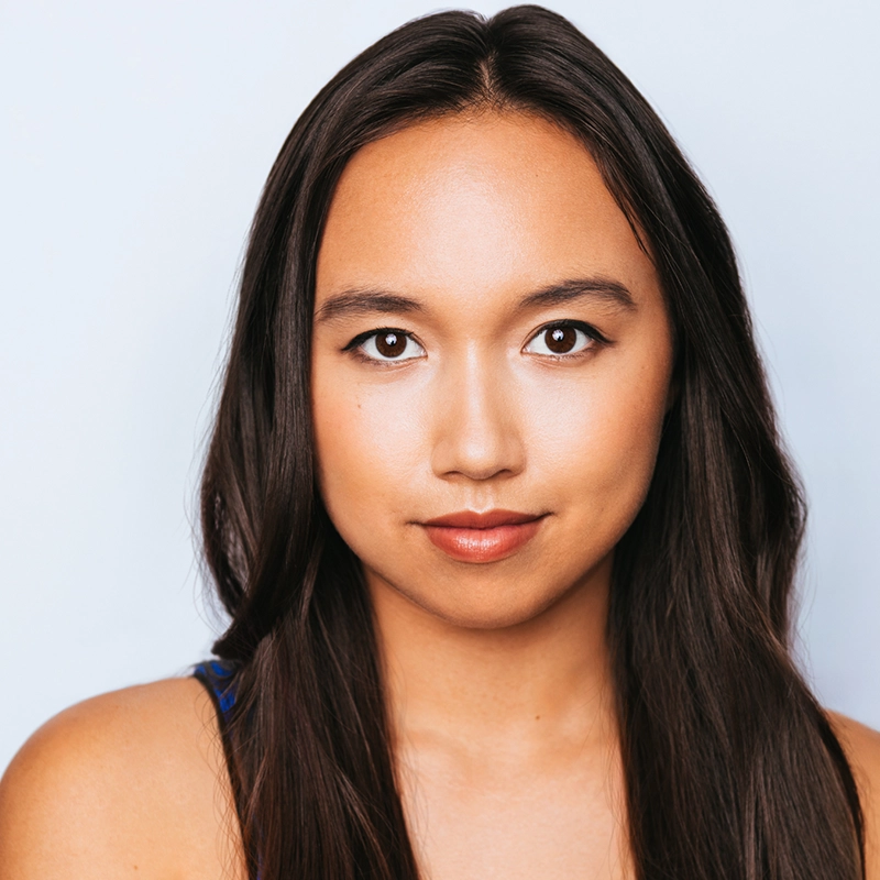 A young woman with long, straight dark brown hair and smooth medium-toned skin looks directly at the camera, wearing natural makeup with subtle eyeliner and lip color. She has a calm, confident expression and a light blue background behind her.