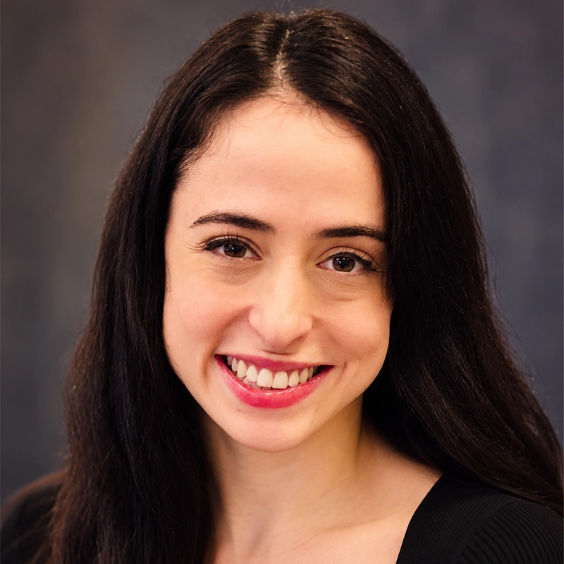 Danny Weller smiles warmly at the camera with her long, dark hair. She wears a black top against a blurred, dark grey background.