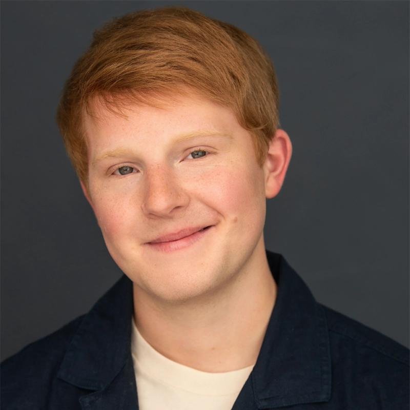 A young person with short, red hair and fair skin smiles gently at the camera. Wearing a dark jacket over a light shirt, Christian Clausnitzer is set against a plain, dark grey background with soft, even lighting highlighting their friendly expression.