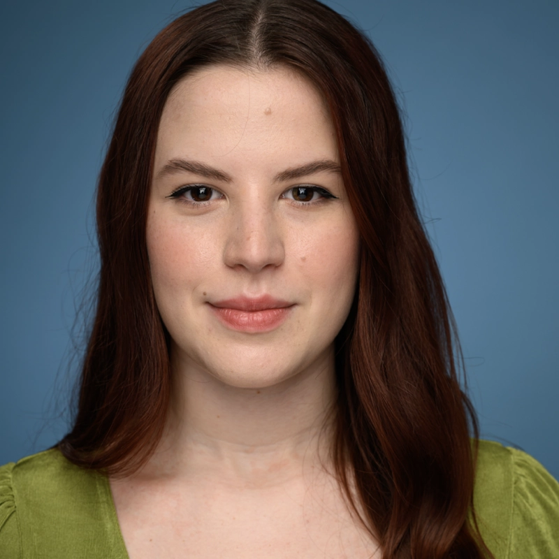 Layla Hope Clarke, a young woman with long, straight auburn hair and fair skin, smiles softly at the camera. She wears a green top and subtle makeup. The plain blue background and even lighting highlight her facial features.