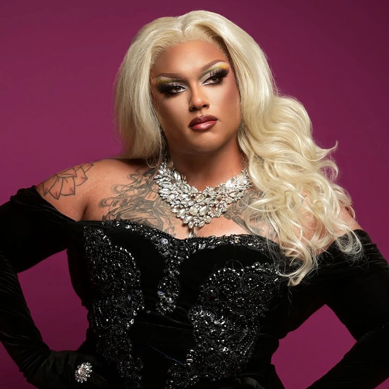 Creatine Price A person with long, wavy platinum-blonde hair poses against a magenta background. They wear dramatic makeup, a jeweled necklace, and a black sequined gown. Tattoos are visible on their left shoulder and chest, creating a striking look as bold as creatine's impact.