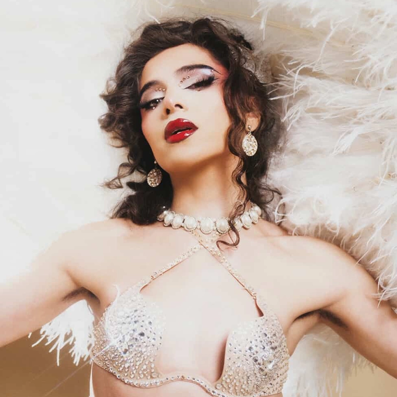 A woman poses dramatically with arms raised, wearing a jeweled bra, pearl necklace, drop earrings, and bold red lipstick. Her curly hair is styled glamorously, and she is surrounded by white feathers, giving a vintage, burlesque-inspired look.