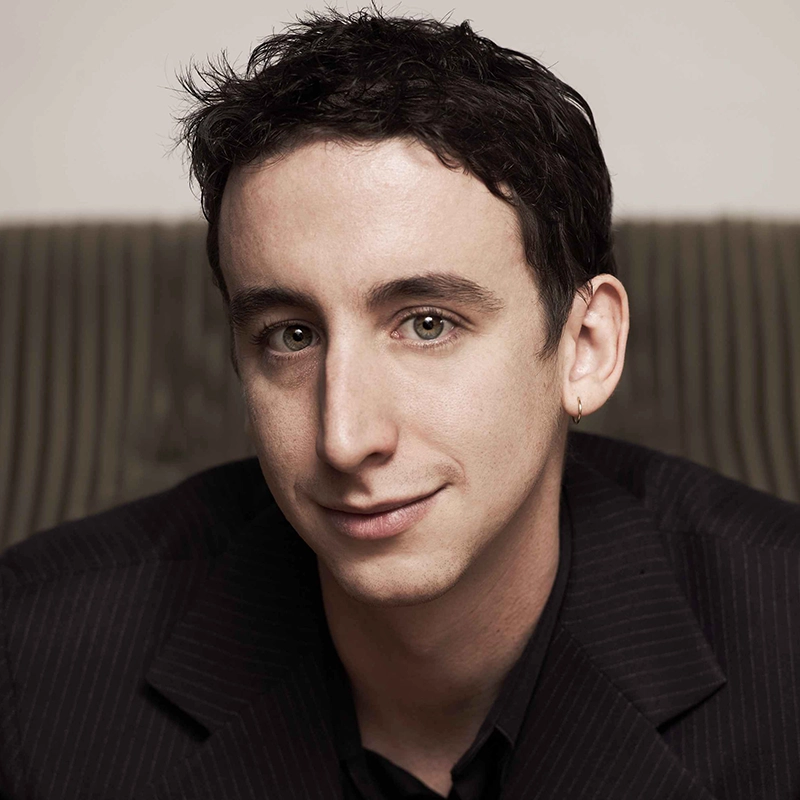 Danny Weller, a young man with short dark hair and a small gold hoop earring in his left ear, looks toward the camera with a slight smile. He wears a dark, pinstriped blazer over a black shirt against a plain background.