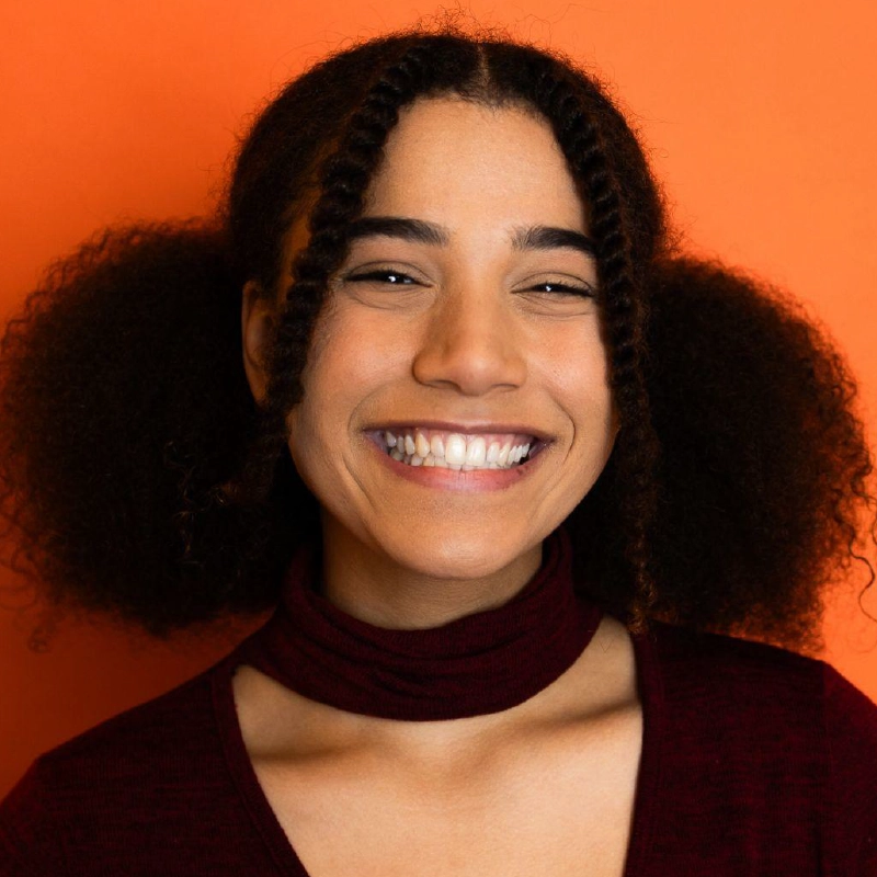 A young woman with medium brown skin and curly hair styled in two puffs and two twisted strands at the front, smiles widely. She wears a burgundy top with a mock neck against a bright orange background.