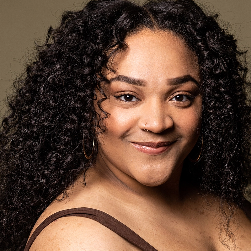 A woman with long, curly black hair smiles gently at the camera. She has medium brown skin, arched brows, and wears hoop earrings and a brown tank top. The softly lit background highlights her face in a portrait inspired by Christian Clausnitzer.