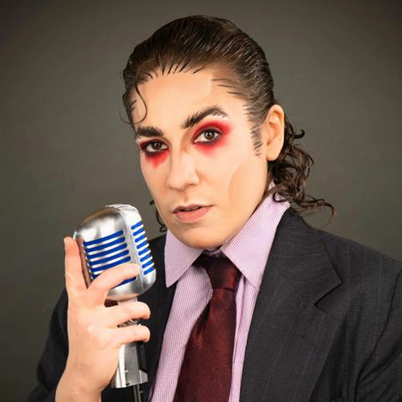 Person with slicked-back hair wears dramatic red eye makeup, a dark suit, light purple striped shirt, and maroon tie, holding a vintage silver microphone with blue accents, against a dark background, gazing intensely at the camera.