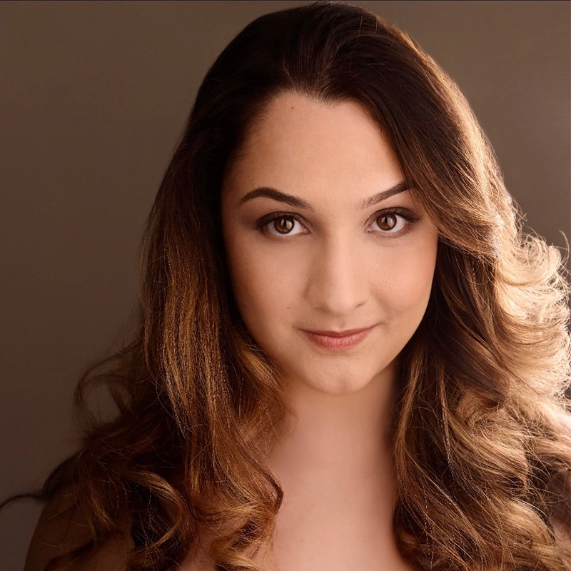 A young woman with long, wavy brown hair and fair skin looks directly at the camera with a soft, confident smile. She has brown eyes and subtle makeup. The softly lit background highlights her face, capturing the poise of Eve Tjoumakaris.