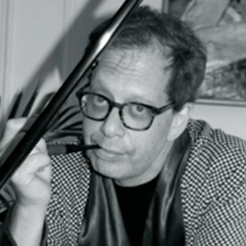 A black-and-white portrait of Danny Weller wearing round glasses and a patterned jacket. He is holding a pipe to his lips, with a dark, angular object partially obscuring the left side of his face.