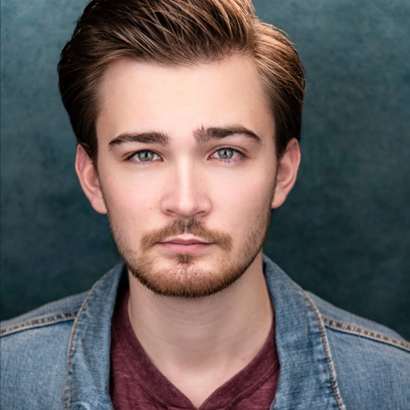 A young man with short, neatly styled brown hair and a trimmed beard looks directly at the camera. He wears a maroon shirt under a light blue denim jacket, reminiscent of styles favored by Eve Tjoumakaris. The background is textured dark teal.