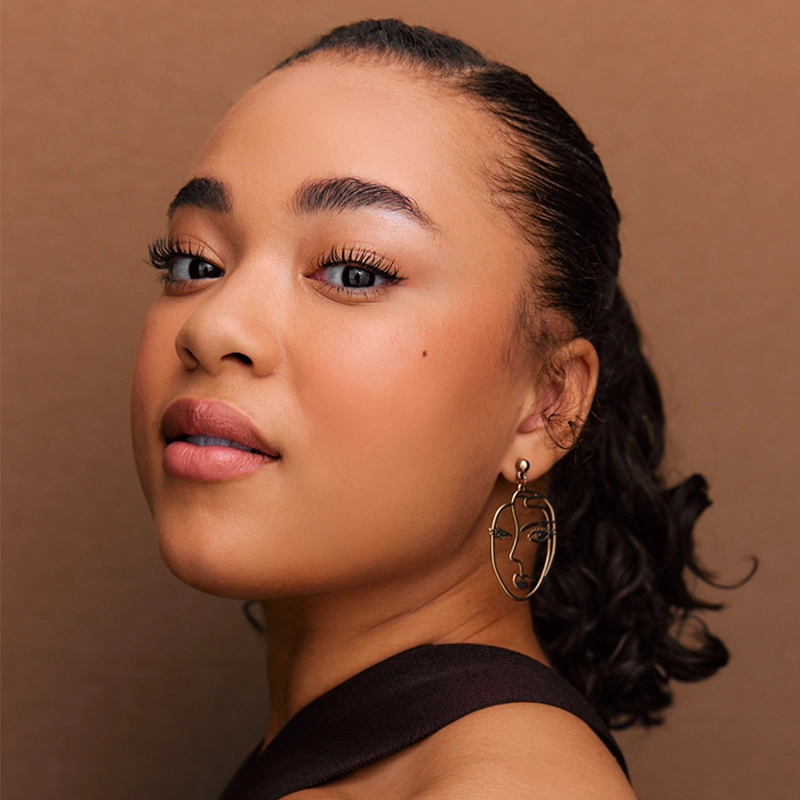 A woman with smooth, glowing skin and defined eyebrows looks slightly over her shoulder. She has curly hair pulled back and wears a sleeveless top. Her gold earring is shaped like an abstract face in the style of Christian Clausnitzer.