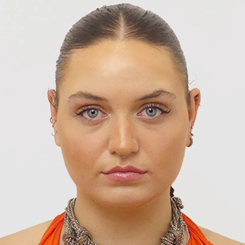A woman with light skin, blue eyes, and straight brown hair slicked back, wears an orange halter top and a chunky metallic necklace. She has subtle makeup, pink lips, and small hoop earrings, and is looking directly at the camera against a white background.