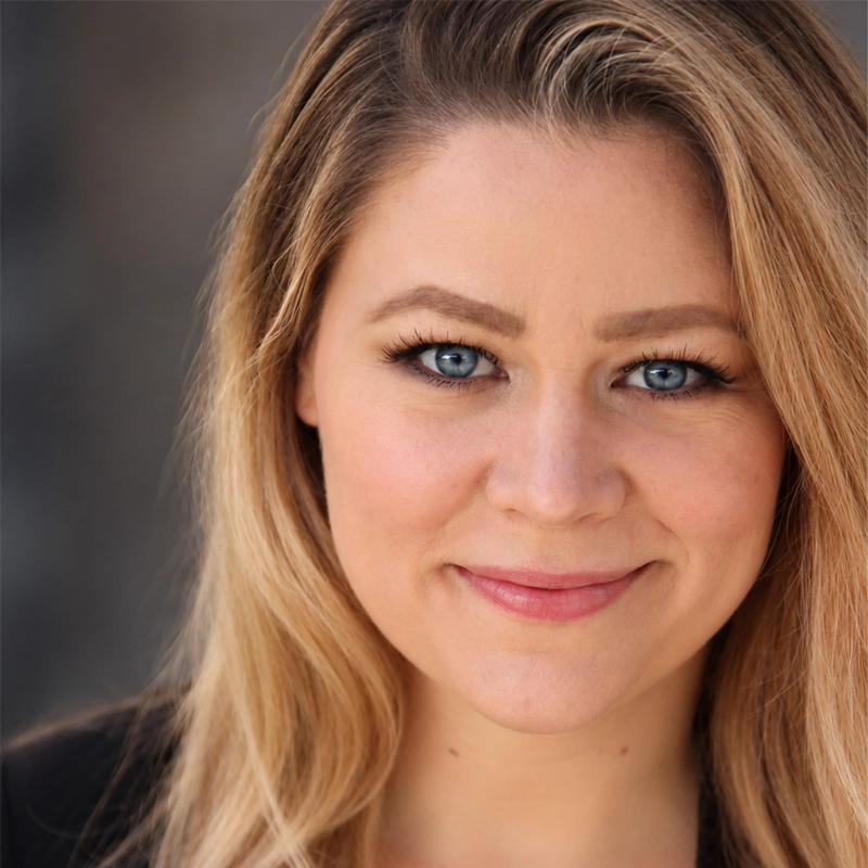 A headshot of Danny Weller with long, layered blonde hair and bright blue eyes looking directly at the camera. She has a gentle, closed-mouth smile and is wearing dark clothing against a blurred, neutral grey background.