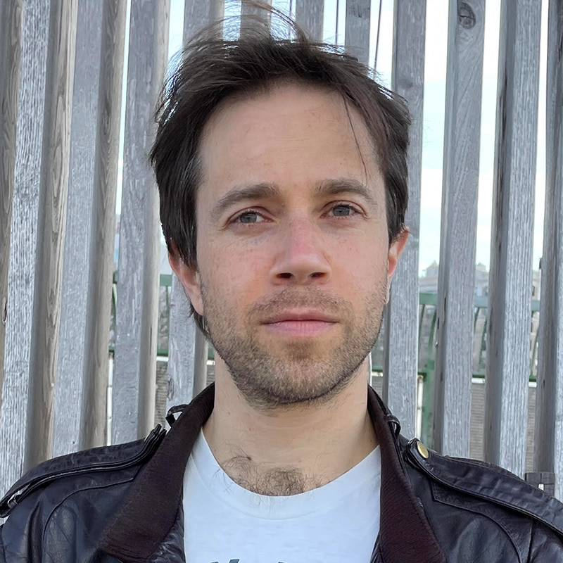 Danny Weller, a man with short brown hair and light stubble, looks directly at the camera. He wears a white t-shirt under a dark brown leather jacket, set against a background of vertical wooden slats.