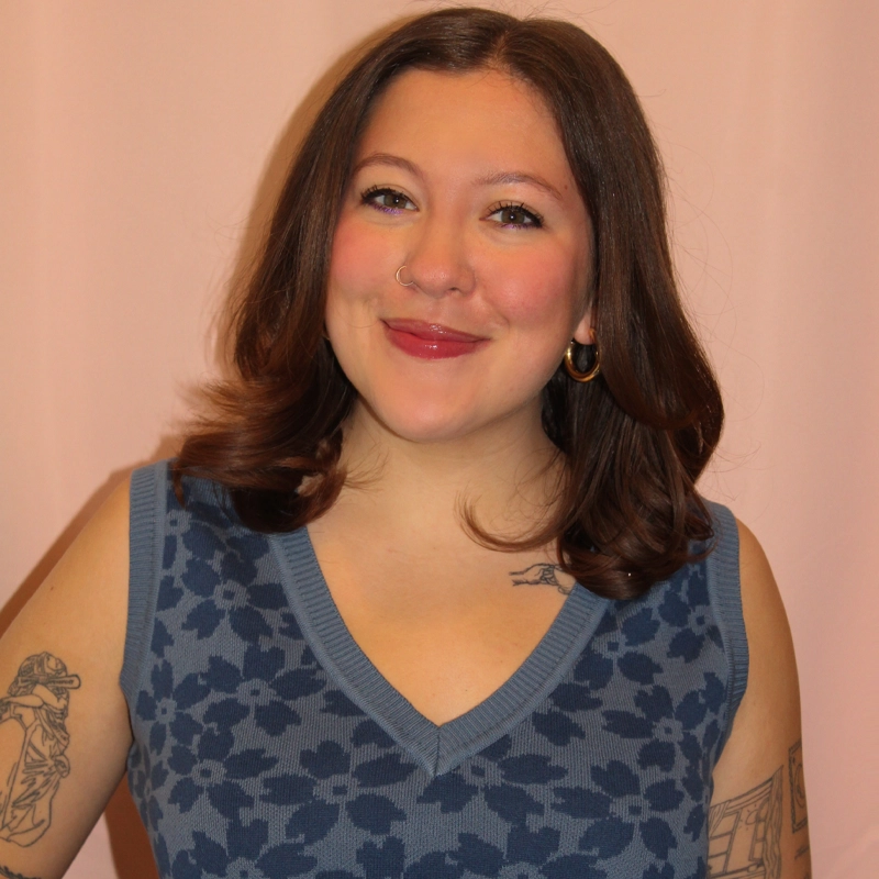 Haley Keizur, with medium-length brown hair, smiles at the camera in a blue patterned sleeveless top. She has tattoos on her arms and chest, wears a nose ring and hoop earrings, and stands against a plain, light pink background.